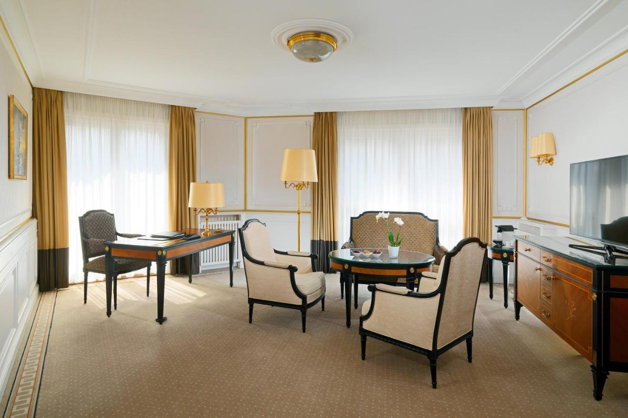 Hotel The Westin Grand Berlino Esterno foto The photo depicts a well-designed room, likely a hotel suite or an upscale living space. It features two large windows covered with light-colored drapes, allowing natural light to fill the room. The flooring appears to be carpeted, contributing to a 