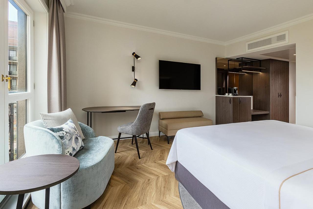 Hotel The Westin Grand Berlino Esterno foto The photo shows a modern hotel room. It features a large bed with white linens, a stylish chair and a small round table to one side. There is a flat-screen television mounted on the wall across from the bed. In the background, there's a kitchenette a