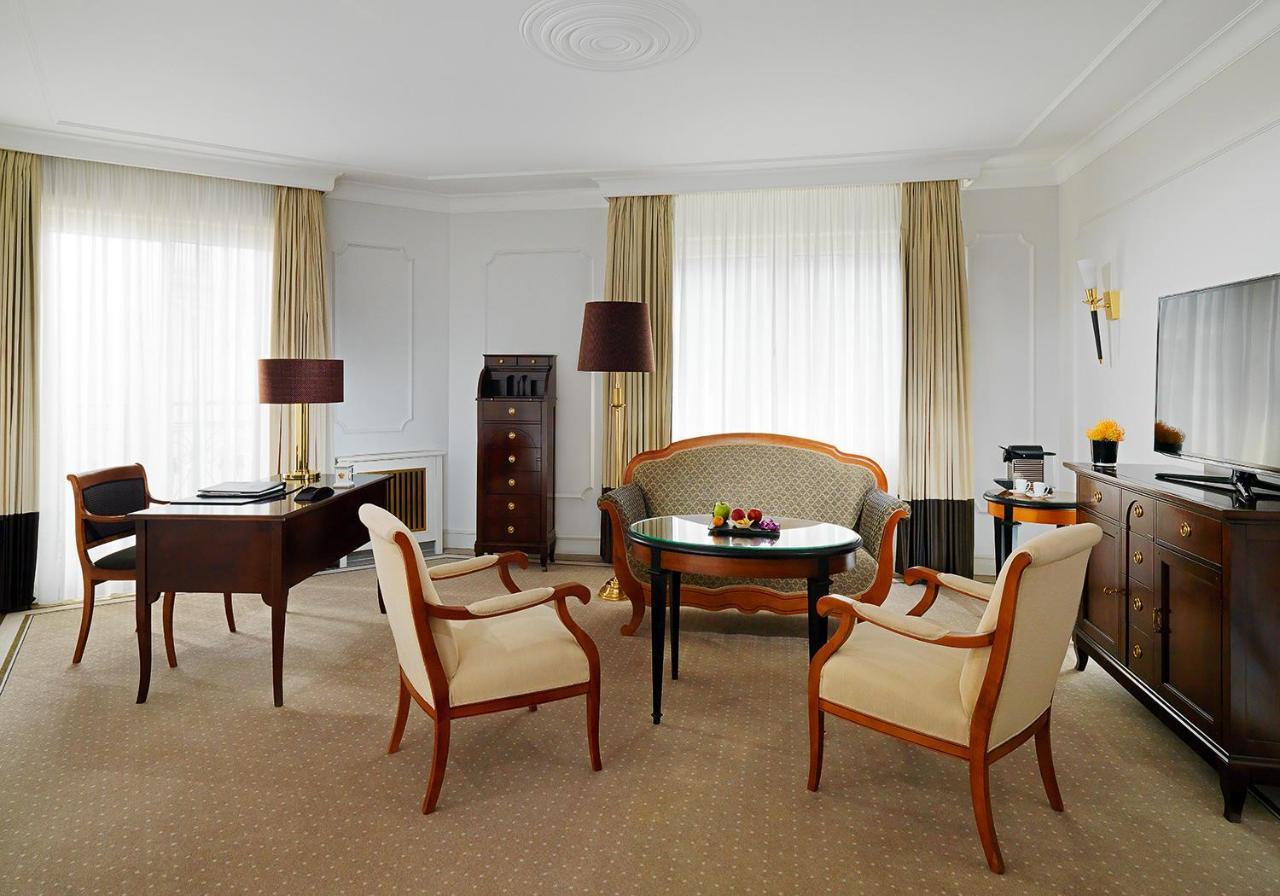 Hotel The Westin Grand Berlino Esterno foto The photo shows a stylish hotel room interior. It features a cozy sitting area with a curved sofa and two light-colored upholstered chairs, all arranged around a small circular table. A dark wooden desk with a lamp and a vintage-style wooden cabinet 