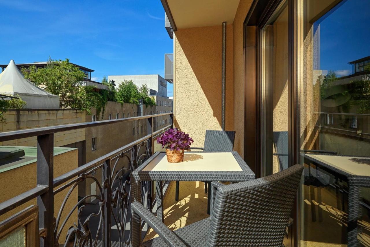 Hotel The Westin Grand Berlino Esterno foto The photo depicts a balcony featuring a small table and two chairs. The table is set with a potted purple flower, adding a touch of decoration. The balcony has a wrought iron railing and glass walls, allowing for outdoor views. The background shows g