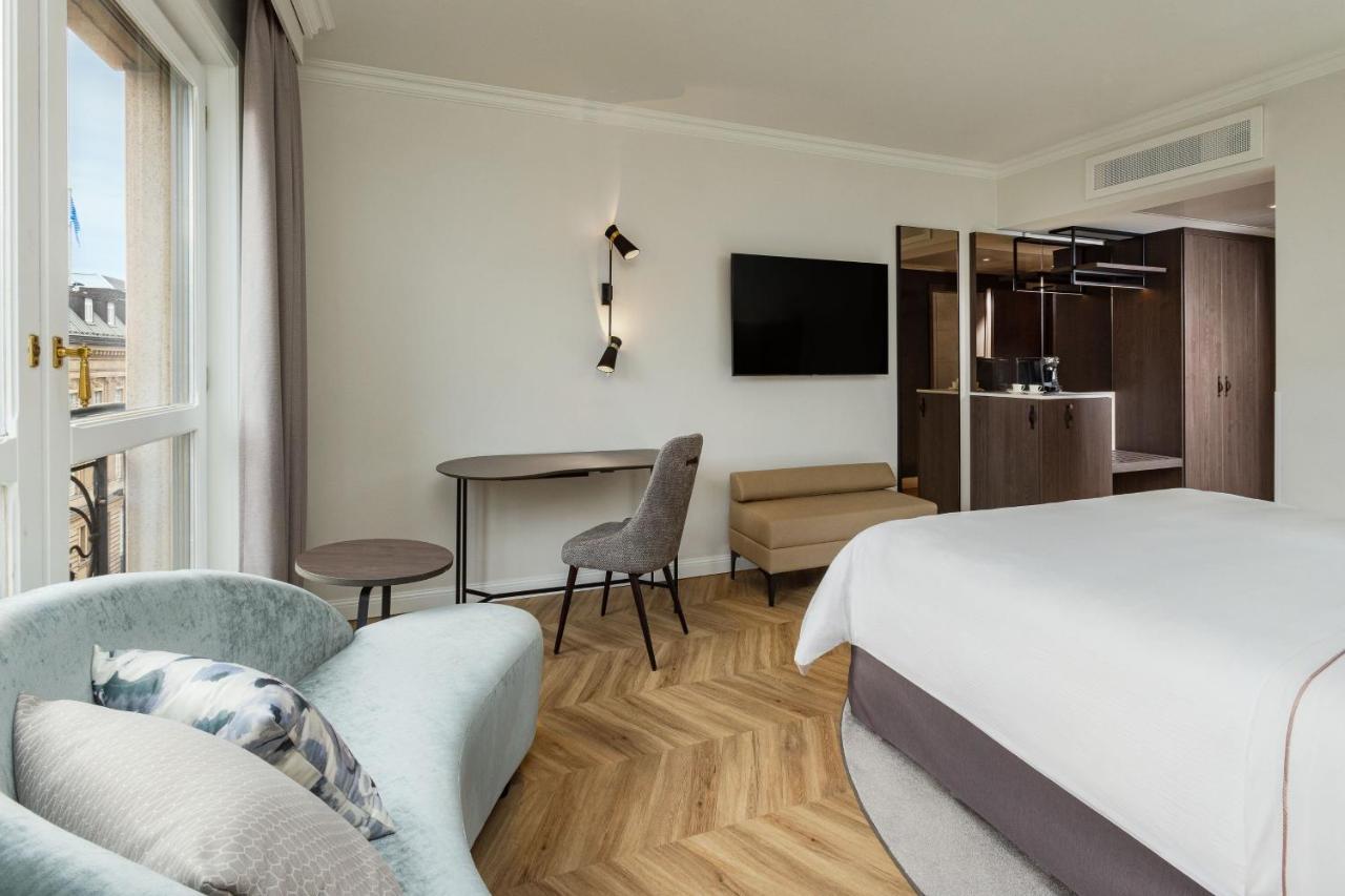 Hotel The Westin Grand Berlino Esterno foto The photo shows a modern hotel room featuring a stylish and contemporary design. There is a comfortable bed with a light-colored bedding set, and a small sofa with a decorative pillow nearby. A round table and a gray chair are positioned in the room,