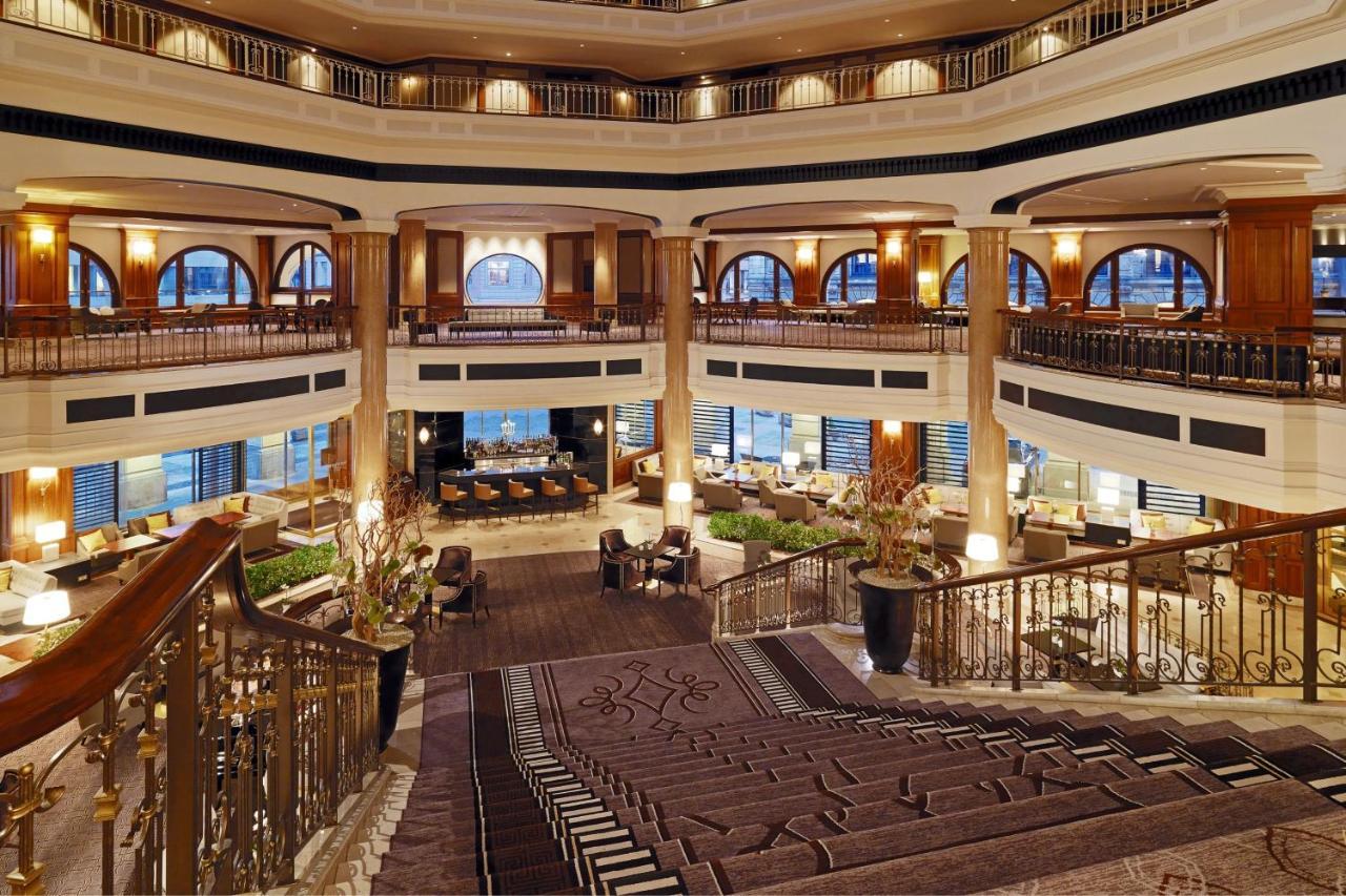 Hotel The Westin Grand Berlino Esterno foto The photo depicts the interior of a luxurious hotel lobby. It features a grand multi-level design with a sweeping staircase that leads down to a spacious area. The lobby has elegant architectural elements, including large arched windows that allow na