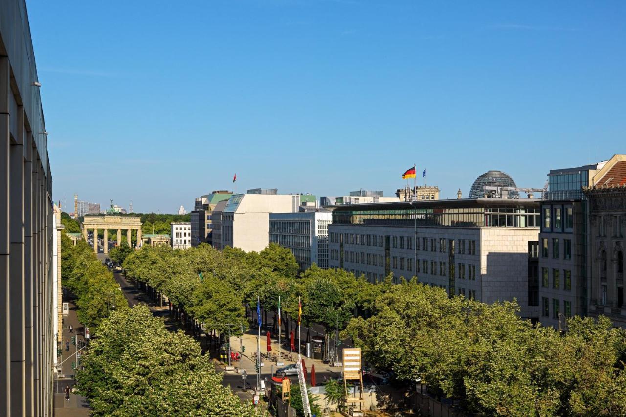 Hotel The Westin Grand Berlino Esterno foto The photo shows a view of a cityscape with a clear blue sky. In the foreground, there is a broad avenue lined with trees. On one side of the avenue, you can see modern buildings, some of which have flags flying. Notably, there's a flag of Germany pro