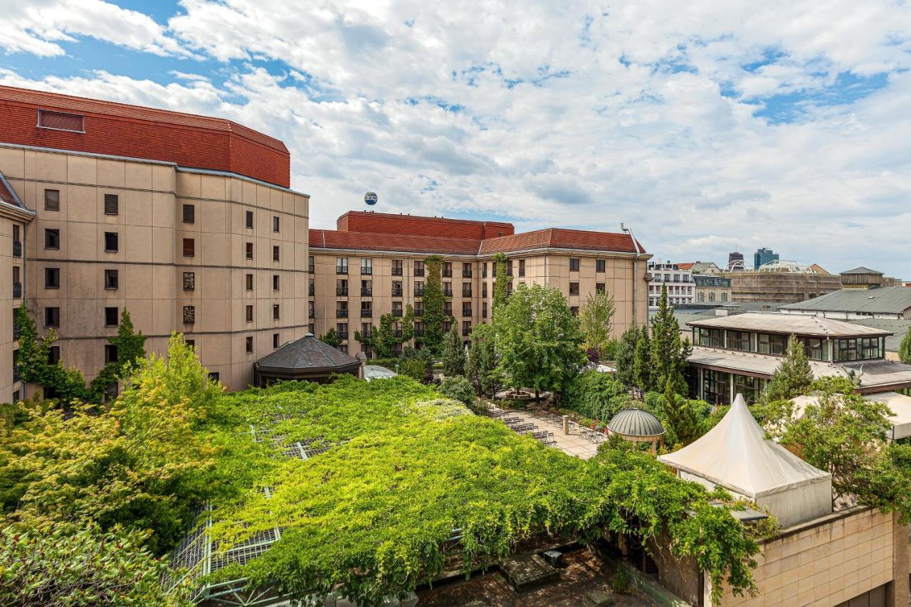 Hotel The Westin Grand Berlino Esterno foto The photo shows a view of a building complex, likely a hotel or similar establishment, with a prominent red roof. In the foreground, there is a lush green area with a lot of vegetation, including trees and vines. There appears to be a tent structure 
