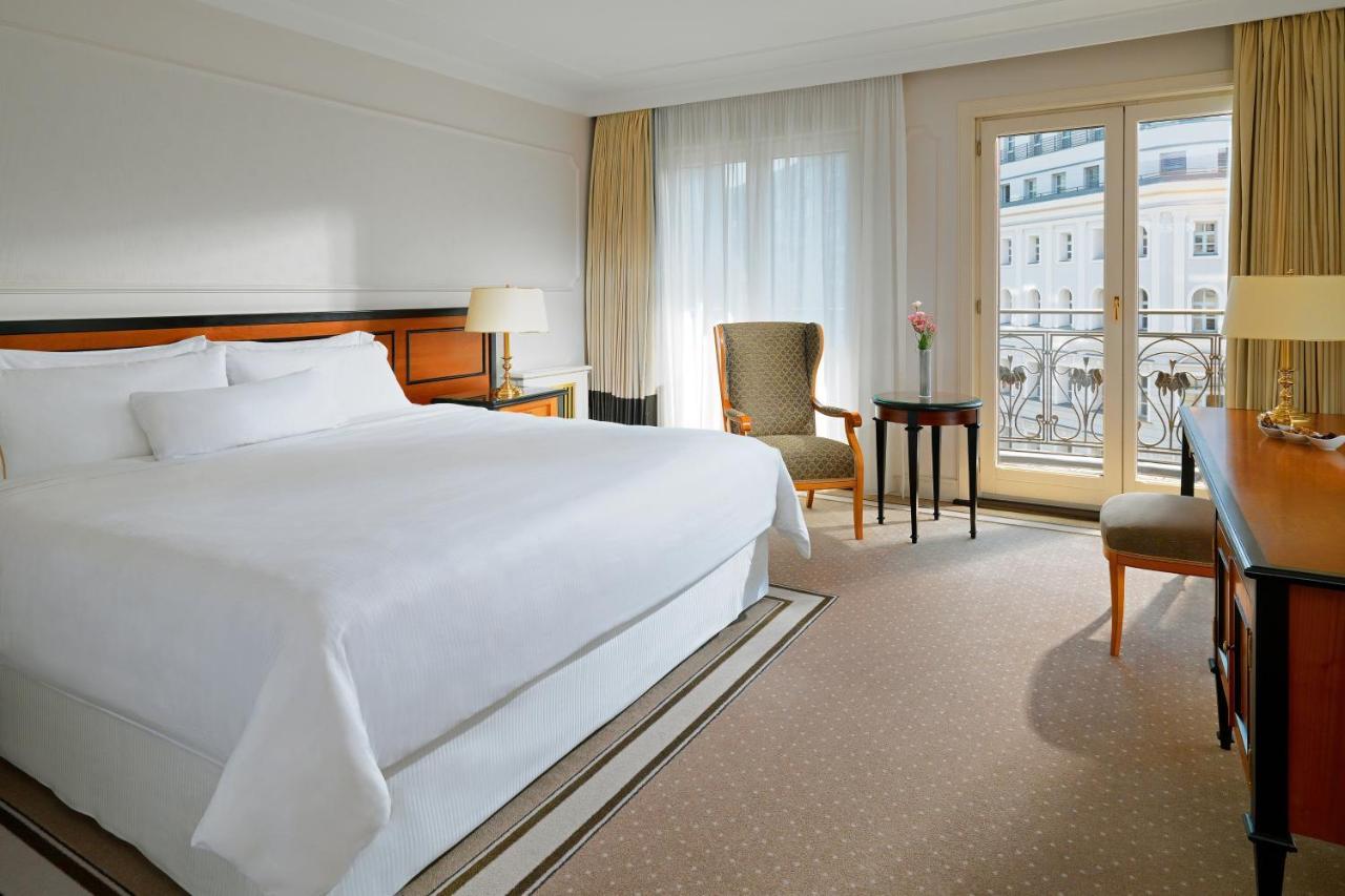 Hotel The Westin Grand Berlino Esterno foto The photo shows a hotel room featuring a large bed with white linens, a bedside lamp, and a small armchair. There is a table next to the chair, and large windows with sheer curtains allow natural light to fill the space. The decor is simple and elega