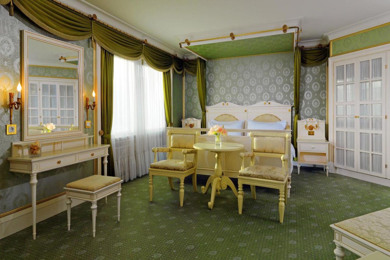Hotel The Westin Grand Berlino Esterno foto The photo shows a beautifully decorated hotel room with a classic and elegant design. The room features a large bed with an ornate headboard, flanked by bedside tables. There are two upholstered chairs and a small round table in the center, providing