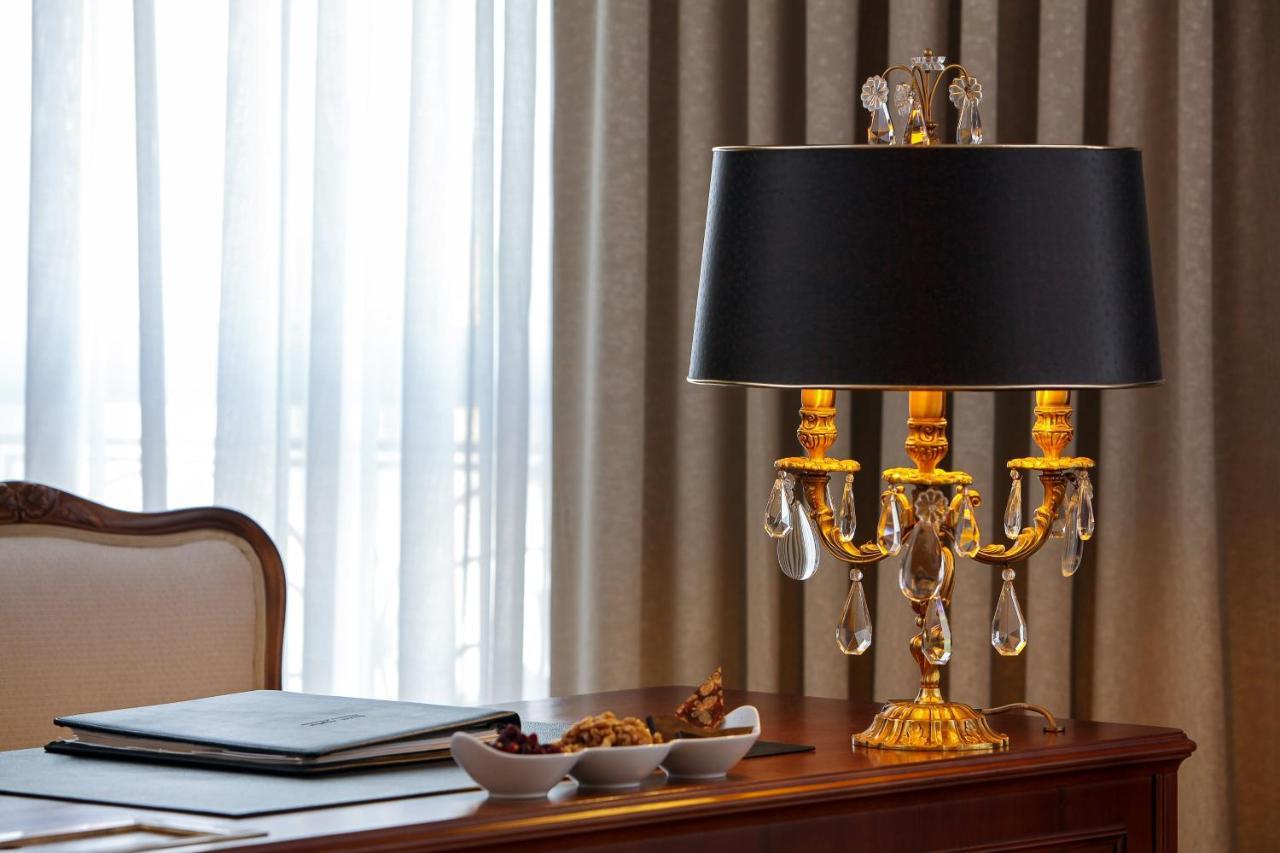 Hotel The Westin Grand Berlino Esterno foto The photo shows a stylish desk lamp placed on a wooden desk. The lamp features an ornate base with a golden finish and decorative elements, including glass or crystal accents that reflect light. It has a black fabric lampshade that contrasts with the