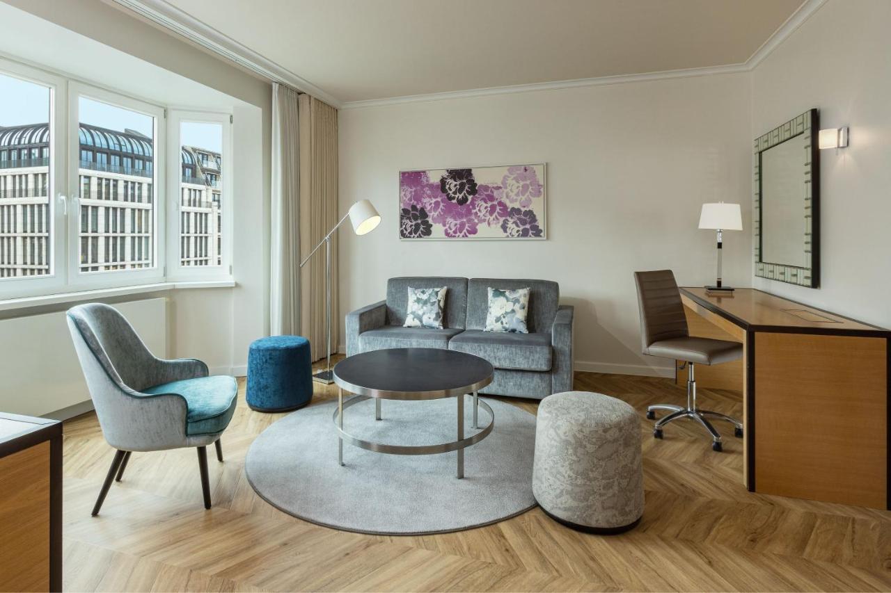 Hotel The Westin Grand Berlino Esterno foto The photo shows a modern and stylish living area. It features a gray sofa with decorative pillows, a round coffee table in the center, and a circular rug underneath. There are two different types of seating: a blue accent chair and a small gray stool