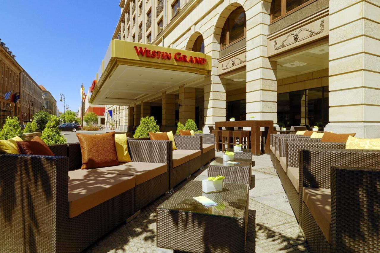 Hotel The Westin Grand Berlino Esterno foto The photo shows an exterior view of a hotel with a prominent sign that reads "Westin Grand." In the foreground, there is a stylish outdoor seating area featuring brown wicker furniture, including couches and coffee tables. The seating is accented wit