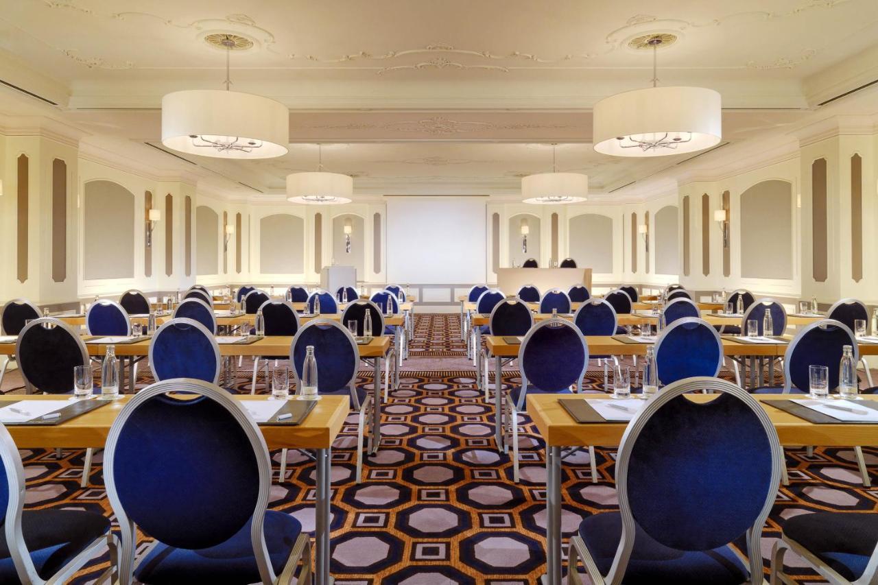 Hotel The Westin Grand Berlino Esterno foto The photo shows a spacious, elegantly designed conference or meeting room. It features multiple rows of tables arranged in a classroom style, with blue upholstered chairs that have circular backs. The floor is adorned with a geometric patterned carpe