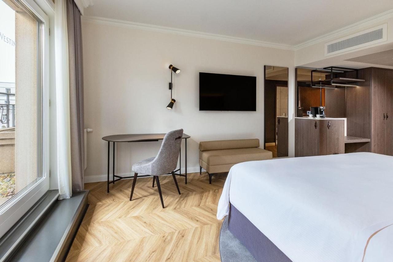 Hotel The Westin Grand Berlino Esterno foto The photo shows a modern hotel room featuring a light-colored interior. There is a bed with crisp white linens, a small seating area with a light-colored sofa, and a round table with a chair. A large flat-screen TV is mounted on the wall, and there i