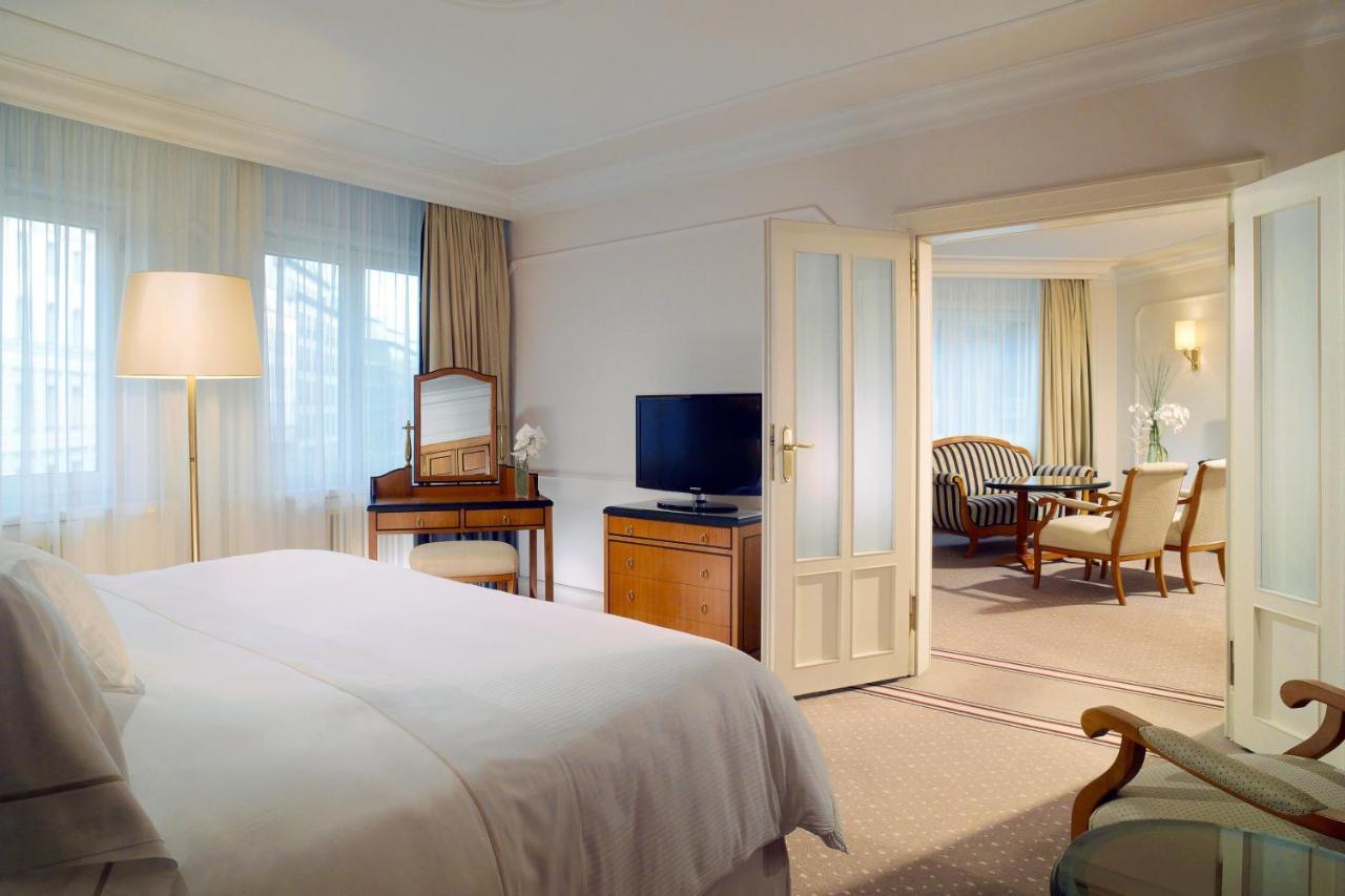 Hotel The Westin Grand Berlino Esterno foto The photo shows a luxurious hotel room. In the foreground, there is a neatly made bed with white linens. To the left, there's a stylish floor lamp illuminating the space. Opposite the bed, there's a television on a wooden cabinet, and a vanity table 