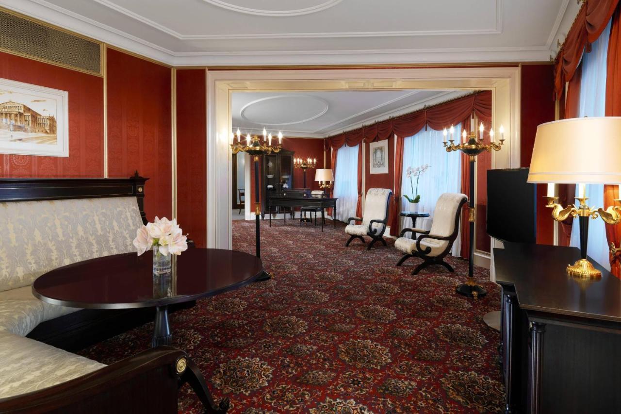 Hotel The Westin Grand Berlino Esterno foto The photo shows an elegantly decorated hotel suite or living space. The interior features rich red wallpaper and a plush patterned carpet, creating a warm and inviting atmosphere. There's a round table with a vase of flowers in the foreground, and tw