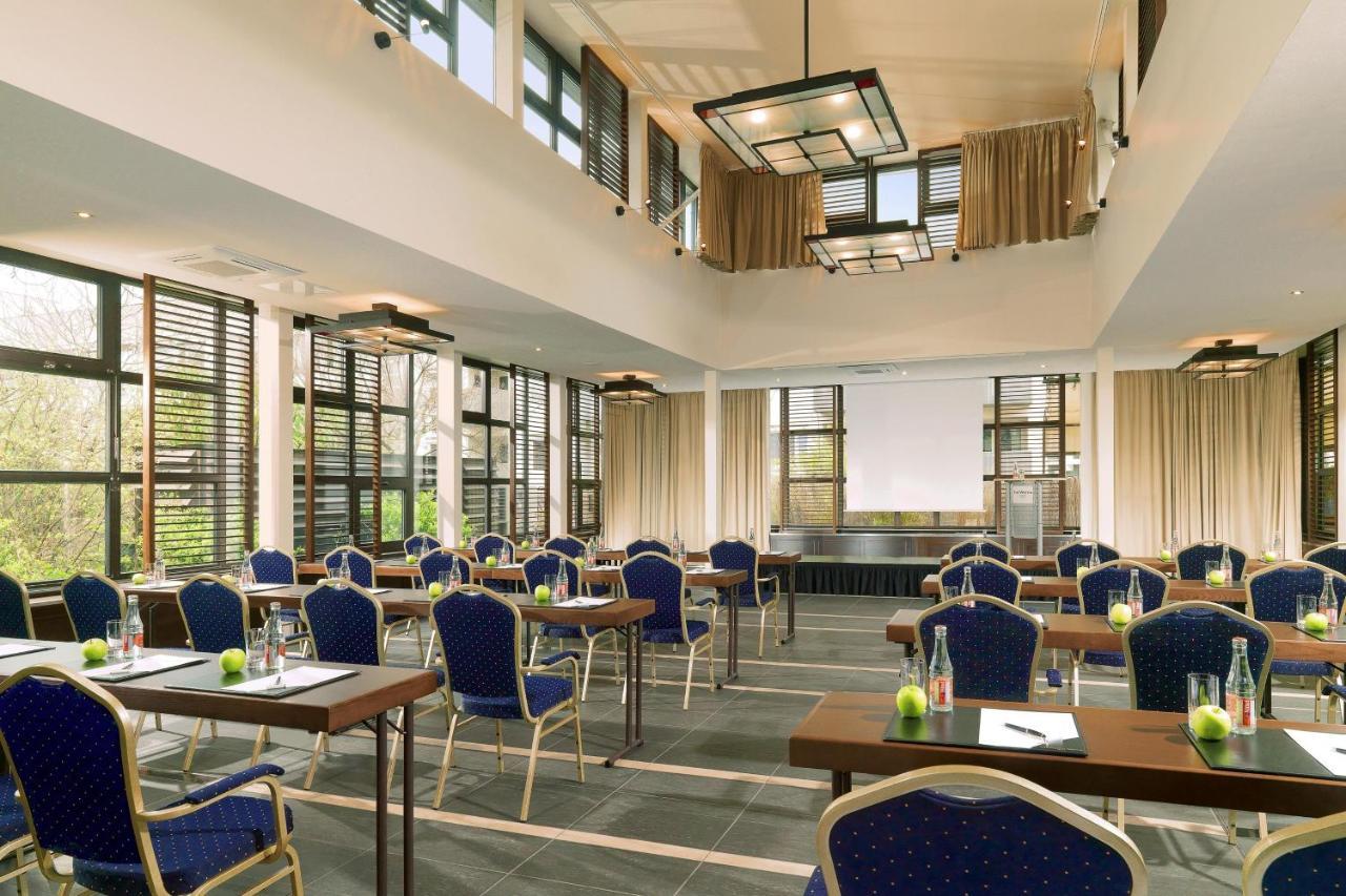 Hotel The Westin Grand Berlino Esterno foto The photo shows a spacious, well-lit conference room designed for meetings or events. It features several rows of wooden tables arranged with blue upholstered chairs. The room has large windows adorned with curtains, allowing natural light to enter. 