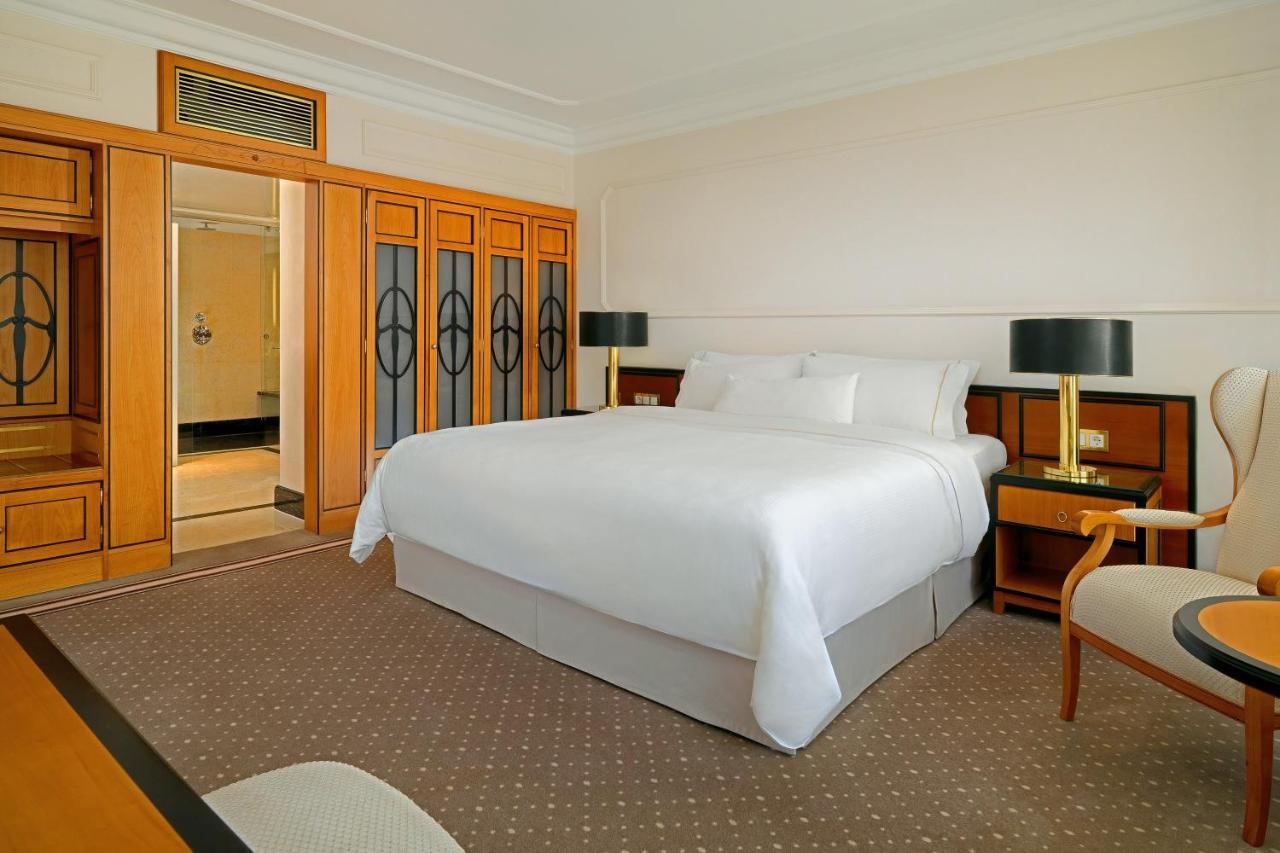 Hotel The Westin Grand Berlino Esterno foto The photo shows a well-furnished hotel room. There is a large, neatly made bed with white bedding in the center. Two stylish bedside lamps are located on either side of the bed. To the left, there is a wooden wardrobe with glass doors. There is also 