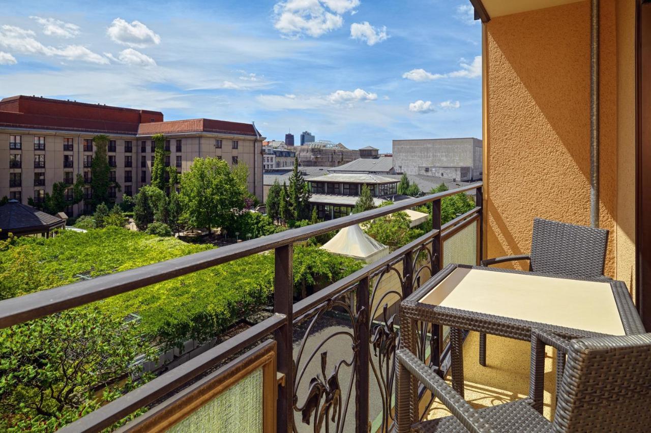 Hotel The Westin Grand Berlino Esterno foto The photo shows a balcony with a view overlooking a vibrant urban scene. There is a railing with decorative design, and a small table with two chairs on the balcony. The area below features greenery and trees, while buildings are visible in the backg