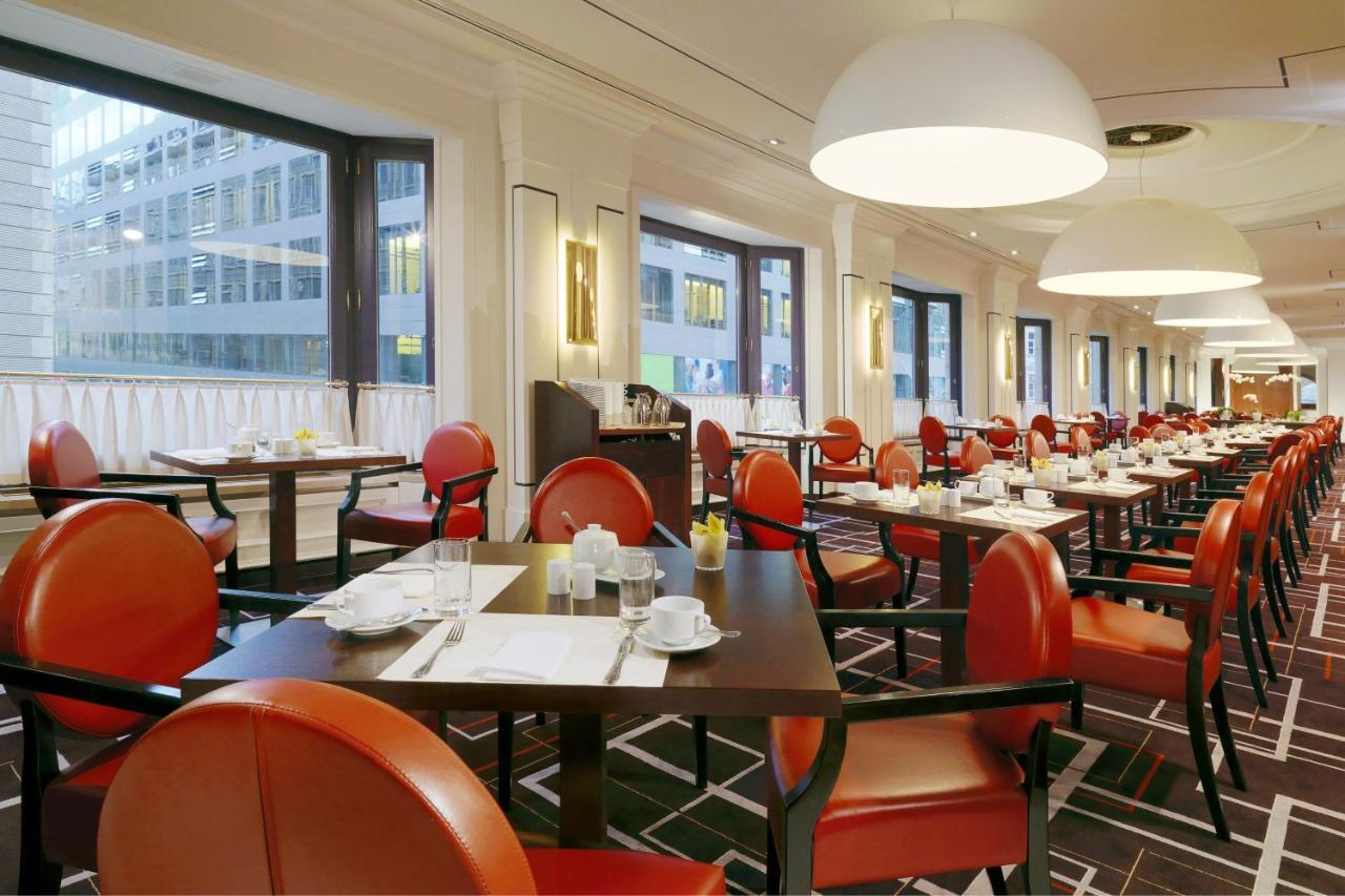 Hotel The Westin Grand Berlino Esterno foto The photo depicts a spacious restaurant interior with a modern design. There are several tables set with white tablecloths and simple dishware, including cups and saucers. The chairs are notable for their orange upholstery, which contrasts with the d