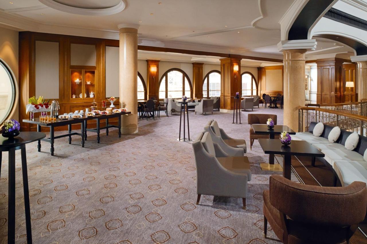Hotel The Westin Grand Berlino Esterno foto The photo shows a spacious and elegantly designed interior of a lounge or reception area. The room features large arched windows that allow natural light to fill the space. There are a mix of comfortable seating arrangements, including plush armchair