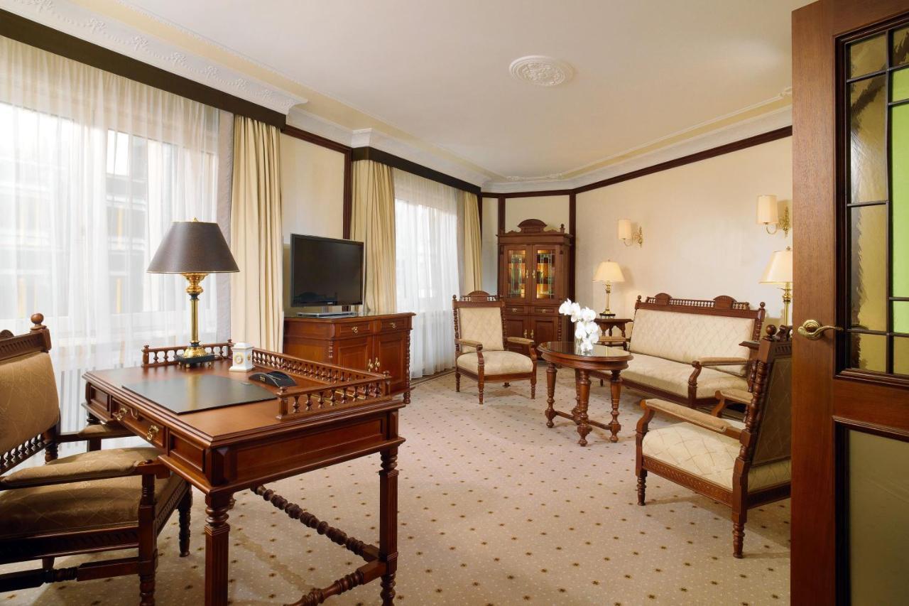 Hotel The Westin Grand Berlino Esterno foto The photo shows an elegant room with a classic design. It features a seating area with upholstered chairs and a sofa, all arranged around a coffee table. A wooden desk with a lamp is positioned nearby, and there is a television on a cabinet. Soft cur