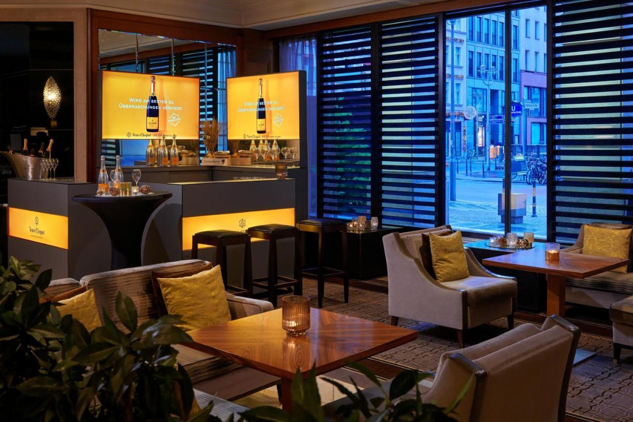 Hotel The Westin Grand Berlino Esterno foto The photo shows a modern bar or lounge area with a contemporary design. There are several tables and comfortable seating arranged around the space, featuring cozy chairs and light-colored cushions. The bar area is well-lit, with stylish displays illu