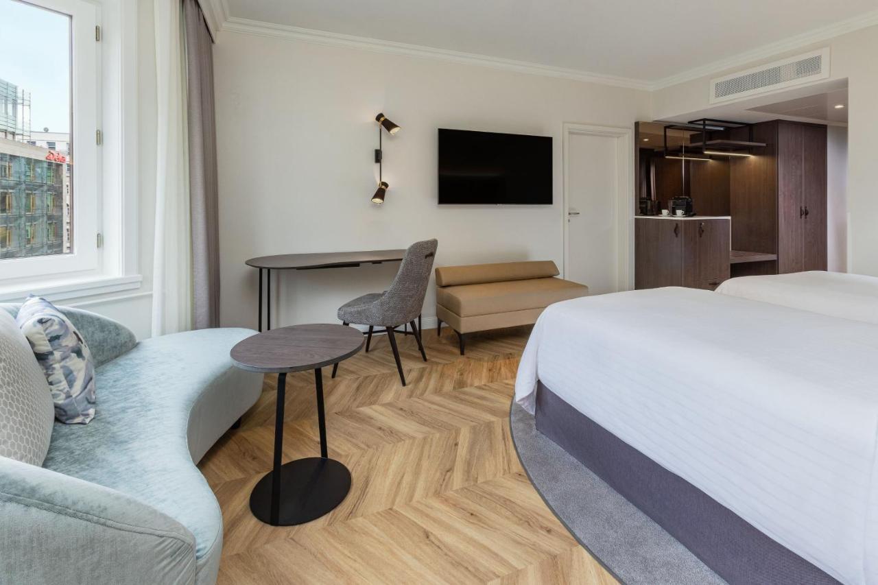Hotel The Westin Grand Berlino Esterno foto The photo shows a modern hotel room. It features a double bed with a light-colored comforter and decorative pillows. There's a small sofa in a soft blue color, accompanied by a round, dark table. A stylish chair is positioned near the table, and a wa