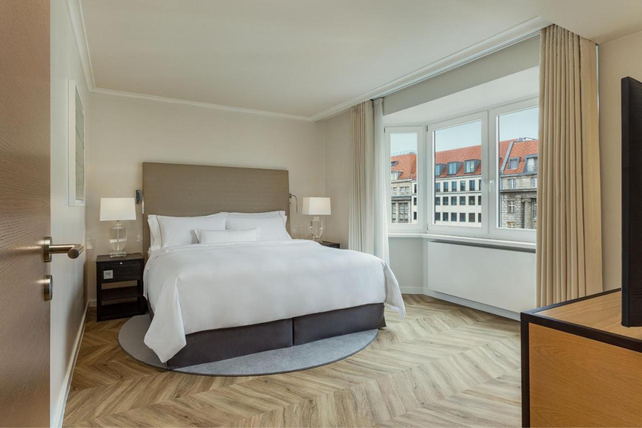 Hotel The Westin Grand Berlino Esterno foto The photo shows a modern, elegantly furnished hotel room. It features a spacious bed with a gray upholstered headboard and crisp white linens. There are two bedside tables with lamps on either side of the bed. The flooring is wood with a herringbone 