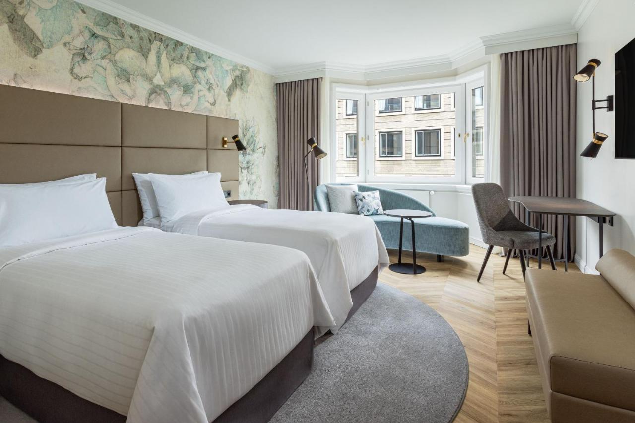 Hotel The Westin Grand Berlino Esterno foto The photo shows a modern hotel room featuring two twin beds with white linens arranged neatly. The walls are adorned with a subtle floral wallpaper, creating a serene atmosphere. There's a small seating area near a large window, which allows for natu