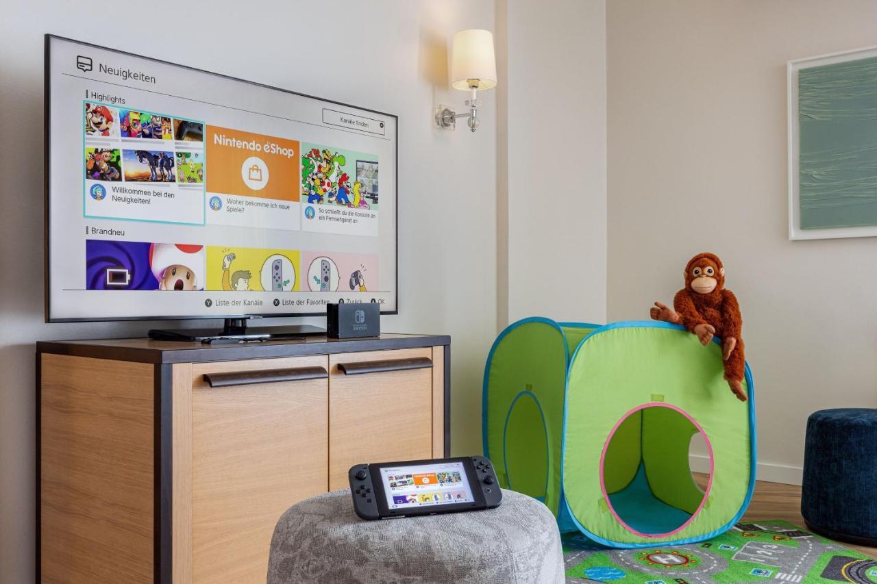 Hotel The Westin Grand Berlino Esterno foto The photo shows a cozy playroom setting. On the left, there is a television mounted on the wall displaying a menu screen, likely for a gaming console. In front of the TV, a gaming console is sitting on a low table, along with a handheld gaming device
