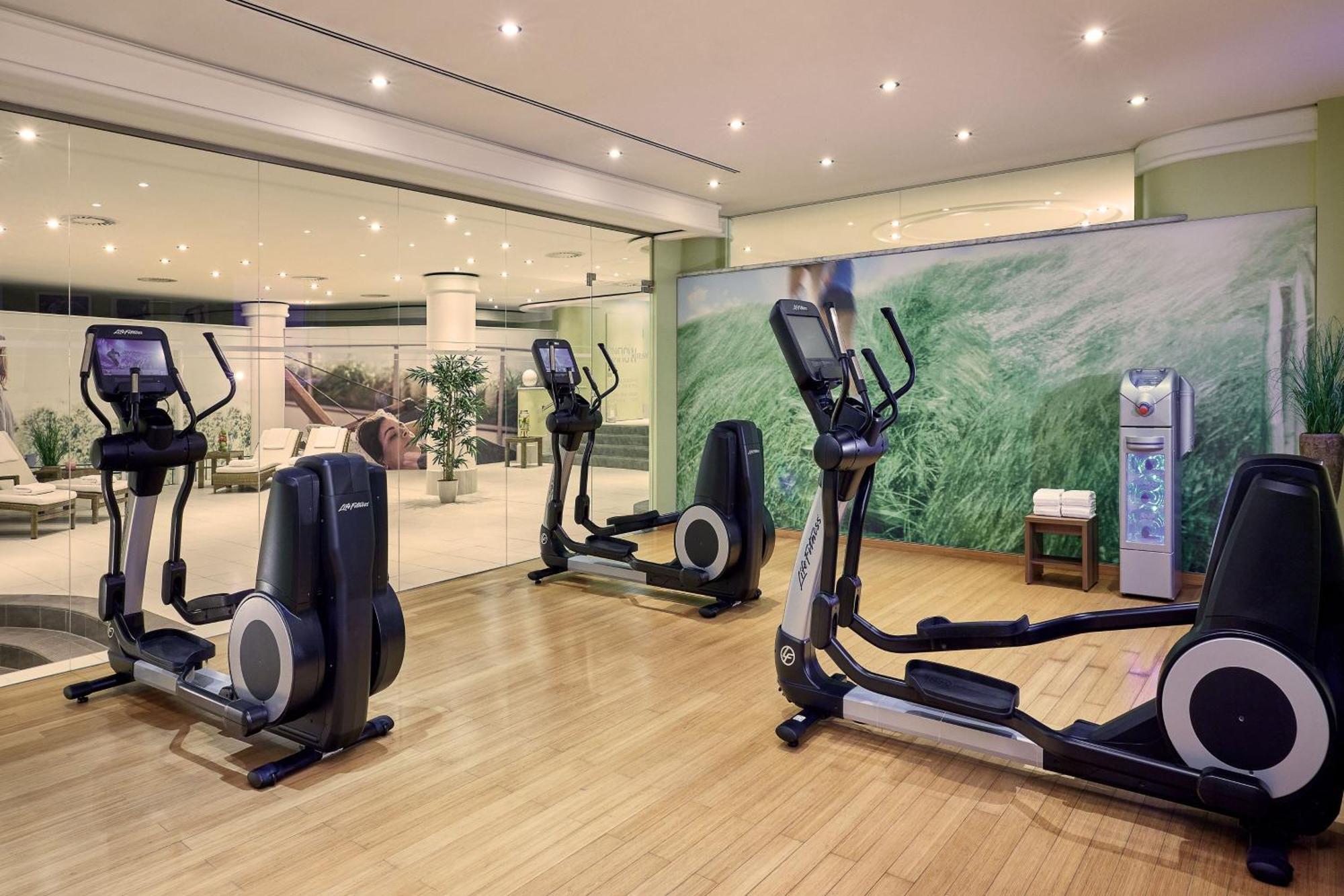 Hotel The Westin Grand Berlino Esterno foto The photo shows a fitness area equipped with several elliptical machines positioned on a hardwood floor. There is a glass wall in the background that allows a view into another room, which appears to be a lounge or common area with plants and seating