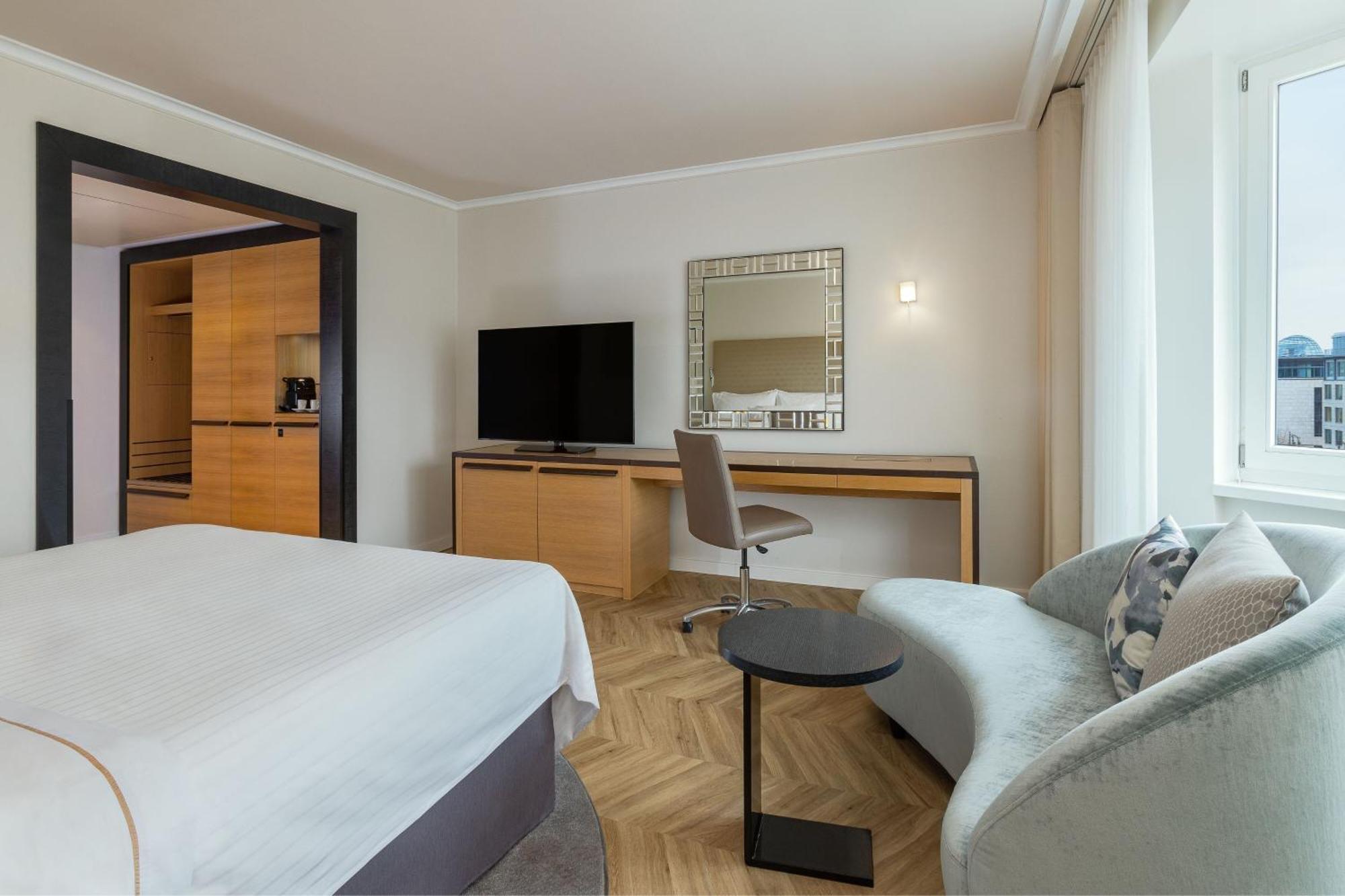 Hotel The Westin Grand Berlino Esterno foto The photo shows a modern hotel room that features several elements:

1. **Bed**: A neatly made bed with a light-colored duvet and white sheets.
2. **Furniture**: There is a small round table next to a contemporary gray sofa, and a desk with a chair p