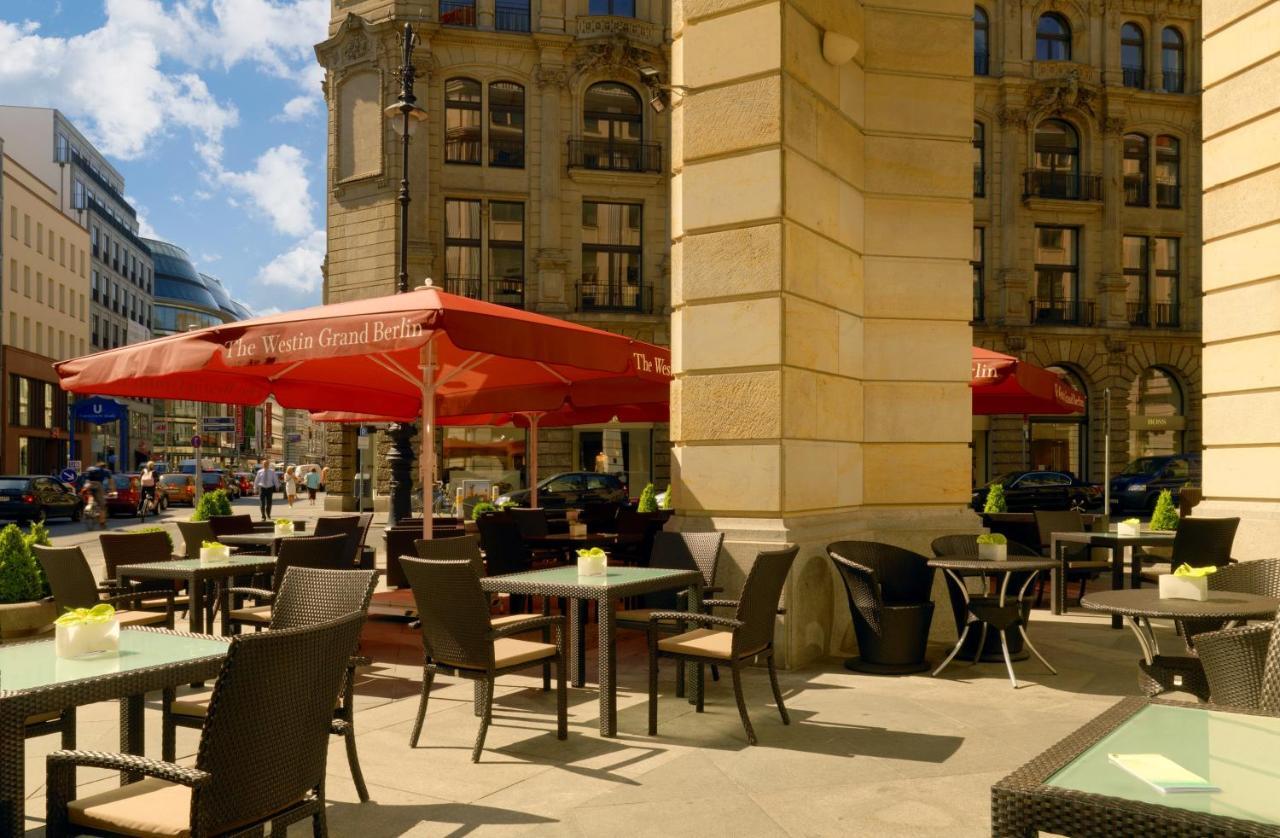 Hotel The Westin Grand Berlino Esterno foto The photo shows an outdoor café area with several tables and chairs arranged under large red umbrellas. There are several potted plants on the tables, adding a touch of greenery. The café is situated in an urban setting, with impressive buildings in 